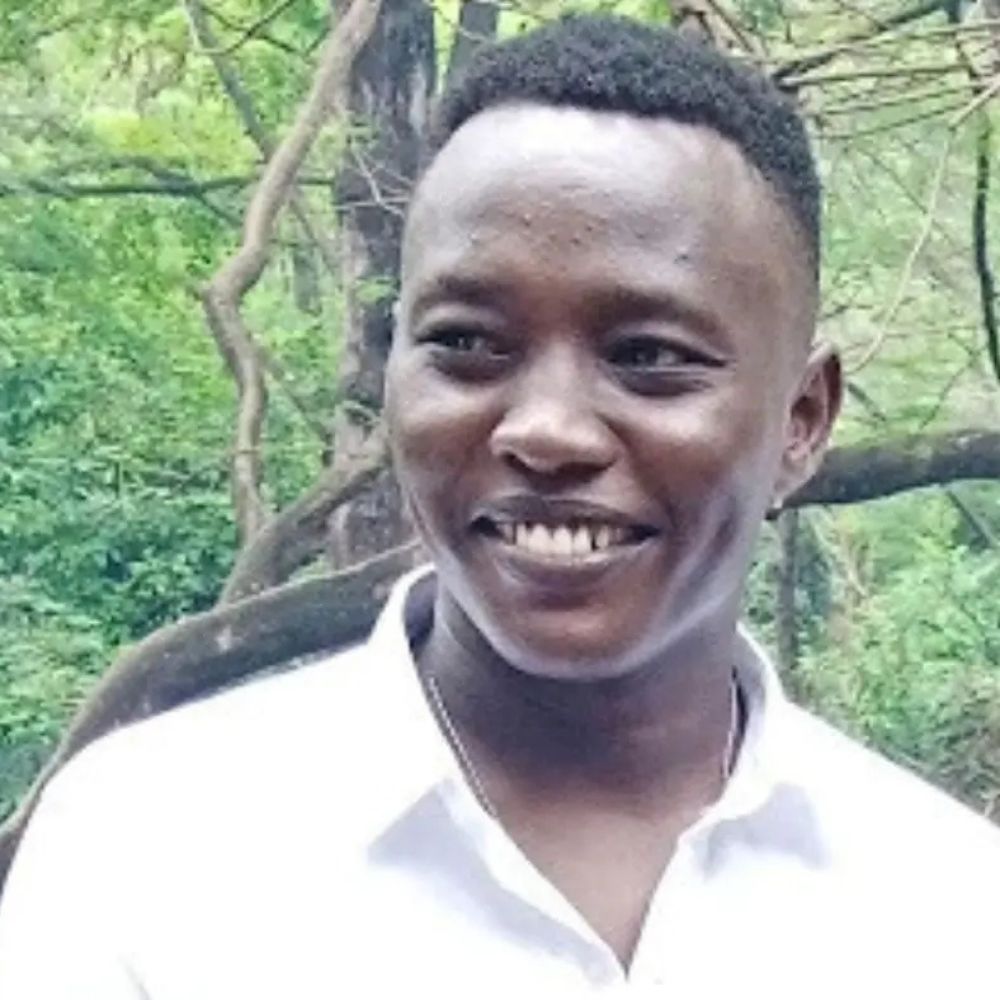 Smiling young man in a white shirt outdoors with greenery in the background.