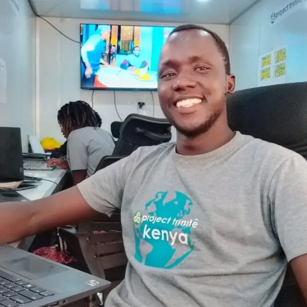 Smiling man in a gray t‑shirt with a teal globe logo sits at a desk with a laptop in a bright office; another person works in the background and a monitor is on the wall.